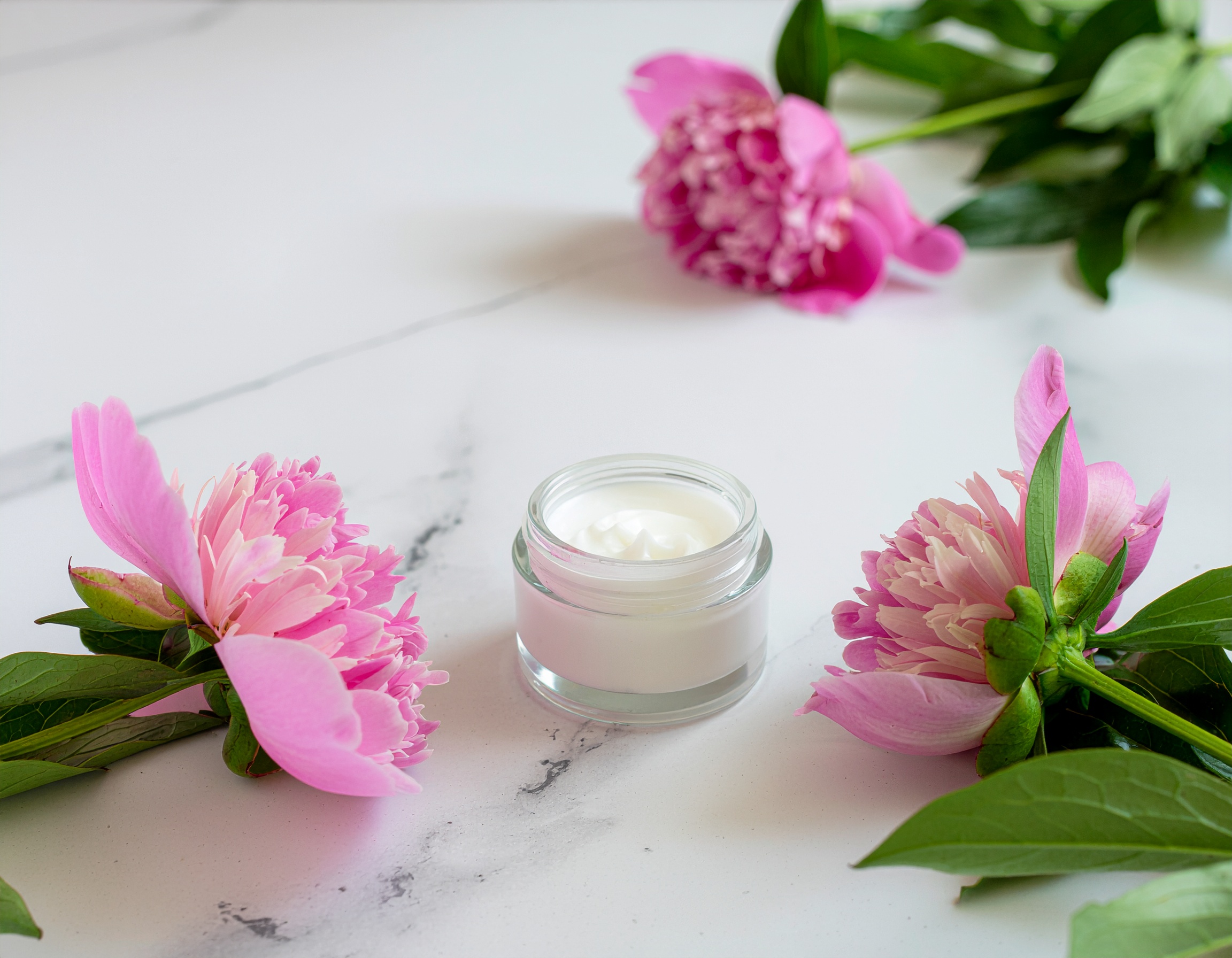 Um frasco de creme hidratante sobre superfície branca, cercado por flores de peônia rosa em um arranjo simétrico. A iluminação suave destaca a textura cremosa do produto, enquanto o fundo claro acentua a delicadeza das pétalas. Composição minimalista transmite sensação de frescor e elegância.